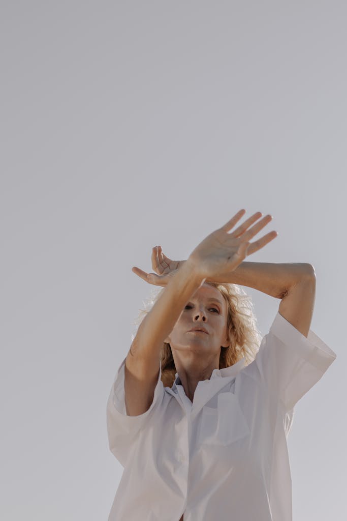 Blonde woman in a white shirt poses elegantly against a clear sky, expressing beauty and grace.